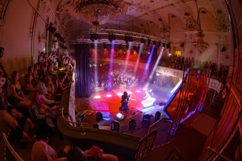 Blick auf eine festliche Aufführung in einem prachtvollen Saal mit einem beleuchteten Podium und einem Publikum auf Balustraden.